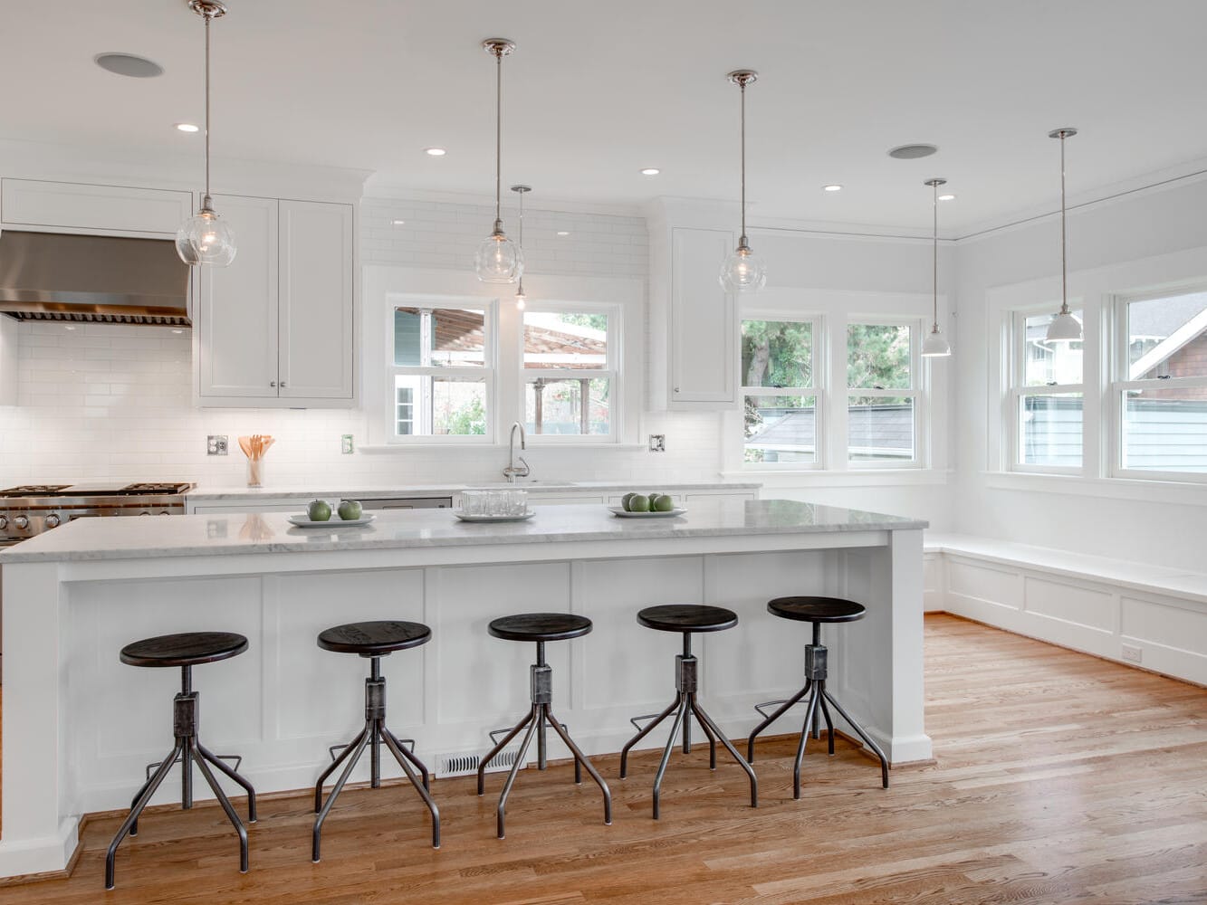 A modern kitchen with a large white island, five black bar stools, stainless steel appliances, and pendant lighting. Wooden floors and multiple windows provide ample natural light.