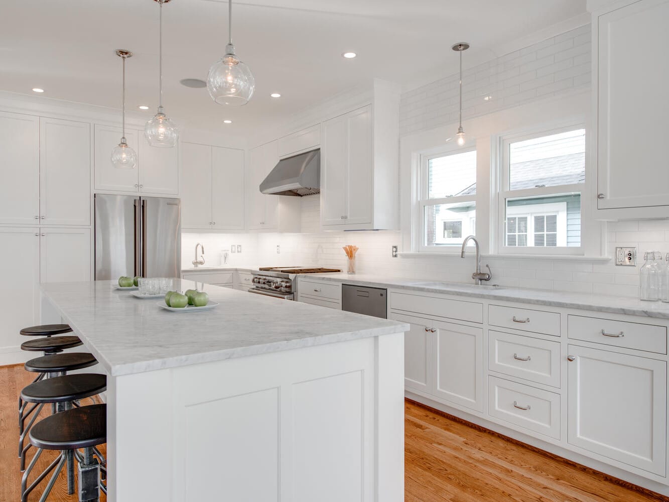 A bright, modern kitchen featuring white cabinetry and marble countertops. Stainless steel appliances are visible, including a refrigerator and stove. Three pendant lights hang above a large island with black stools. The floor is wooden.