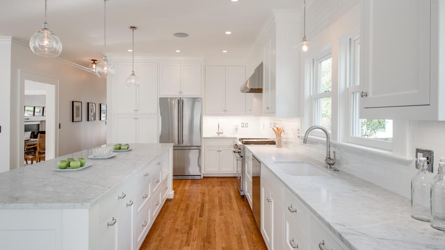 A bright, modern kitchen with white cabinets, marble countertops, and wooden floors. Three pendant lights hang above the island, which has green apples on it. Stainless steel appliances are visible, and a window allows natural light to enter.