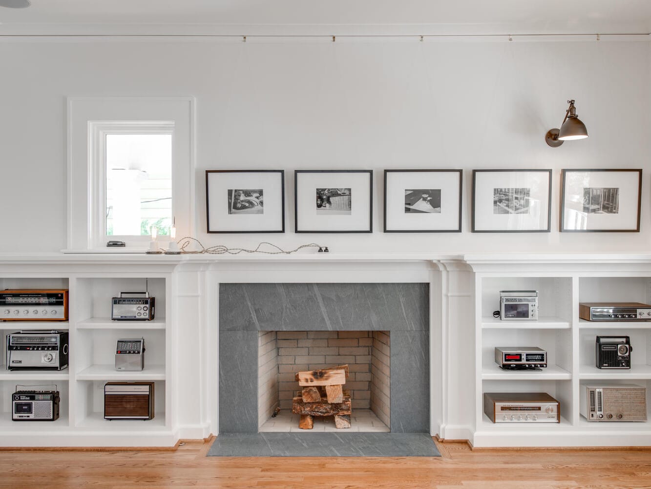 A modern living room features a gray-tiled fireplace with logs inside. Surrounding built-in white shelves display vintage radios. Above the mantel are five black-and-white framed photos. A wall-mounted lamp provides lighting.