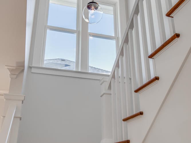 A bright stairway featuring white walls and wooden steps. Three round pendant lights hang from the ceiling. Natural light flows in through a large window with a view of a blue sky.