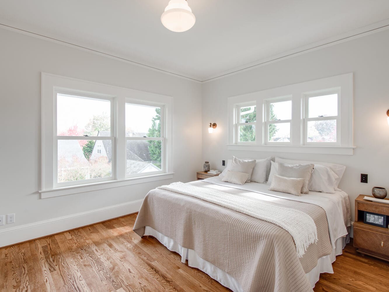 A bright, cozy bedroom with a large bed, white bedding, and wooden side tables. Natural light streams in through four windows, highlighting the hardwood floor and soft neutral wall colors. A pendant light hangs from the ceiling.