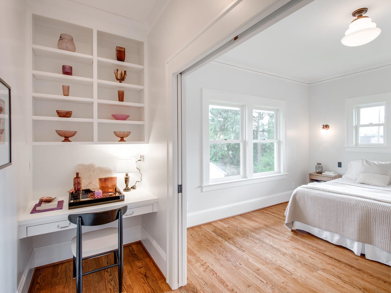 A bright bedroom with a wooden floor features a neatly made bed with beige bedding. On the left, theres a small desk with a chair and decorative shelves displaying pink items. Large windows let in natural light.