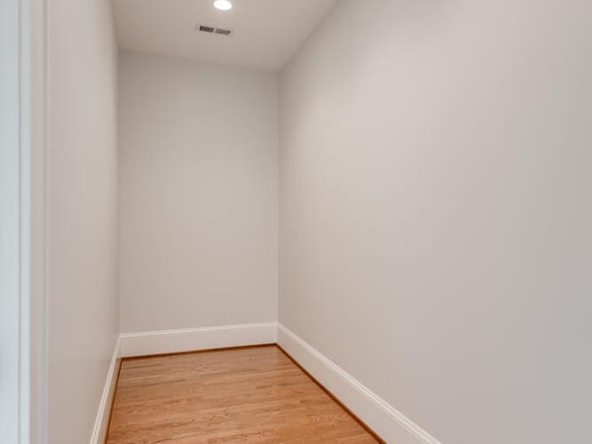 A narrow, empty hallway with light gray walls, wooden flooring, and recessed ceiling lights. The corridor is well-lit and appears clean, with white baseboards lining the bottom of the walls.