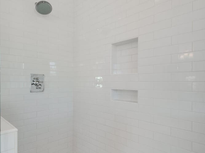 A modern shower with white subway tiles covering the walls, a round shower head, and a temperature control panel. The shower floor has small hexagonal tiles and two built-in shelves in the wall.