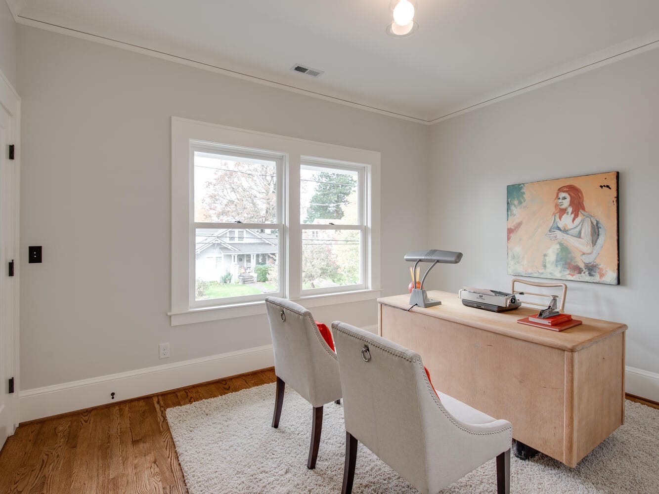 A minimalist home office with a wooden desk, two white chairs, a desk lamp, books, and a record player. A painting hangs on the wall, and a large window offers a view of trees outside. The room has hardwood floors and light-colored walls.