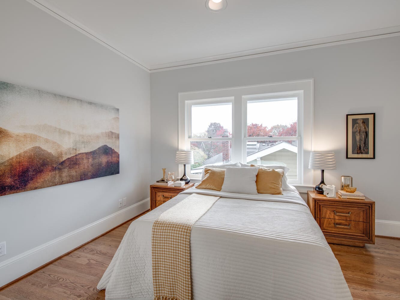 A cozy bedroom with a neatly made bed featuring a beige and white color scheme. Two wooden nightstands flank the bed, holding lamps and decor. A large abstract landscape painting adorns the gray wall, and two windows provide natural light.