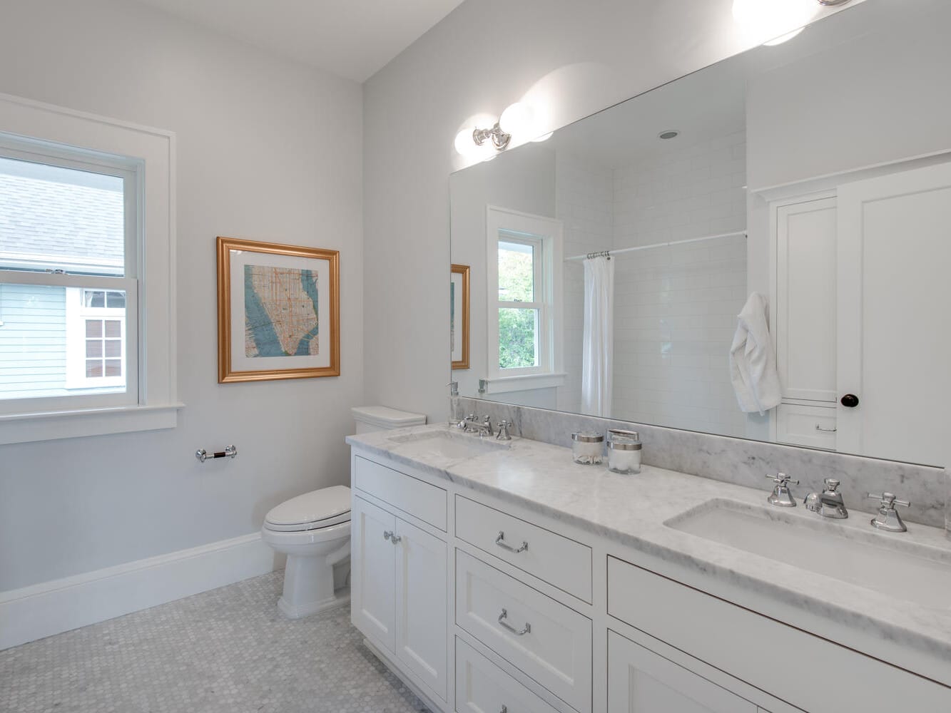 A bright bathroom featuring a large mirror above a white double-sink vanity with marble countertop. There is a bathtub with a shower curtain, a window, and a framed map on the wall near the toilet. The floor is tiled in a light pattern.