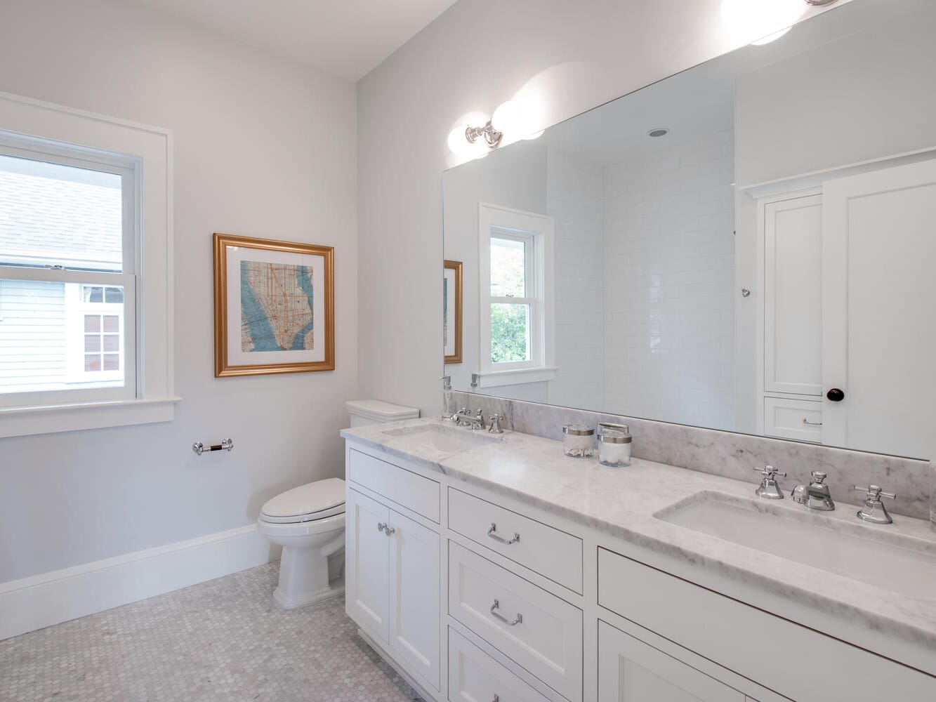 A bright, modern bathroom with a large mirror above a marble-topped vanity. It features dual sinks with chrome fixtures, a framed map on the wall, a toilet, and a window allowing natural light to flood the space.