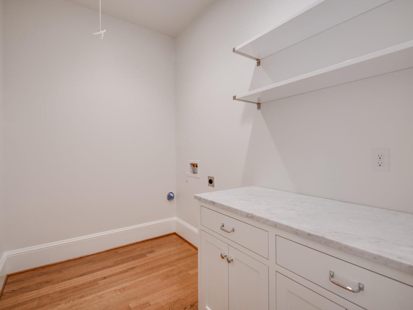 A laundry room with hardwood floors, white walls, and an empty shelf. There is a counter with white cabinets and space for a washer and dryer, along with utility hookups on the wall.