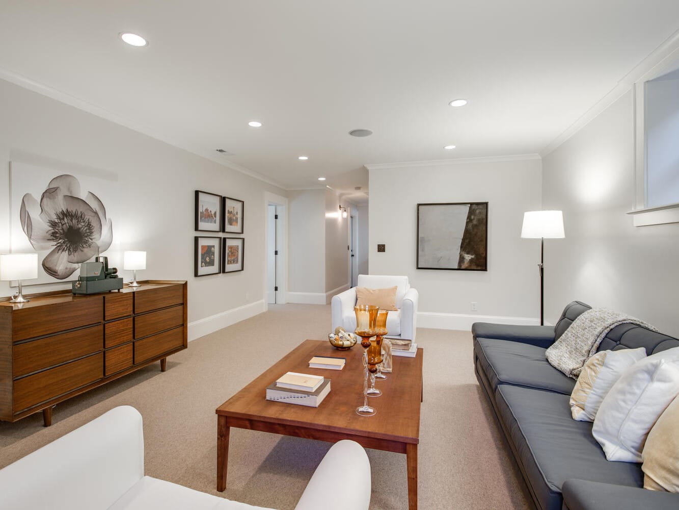 A modern living room with a dark sofa, wooden coffee table, and white armchairs. The room features a wooden sideboard with lamps and artwork on the walls. Ceiling lights illuminate the beige carpeted floor and white walls.