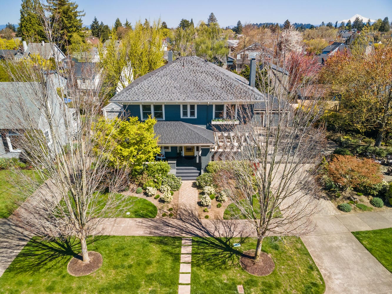 Aerial view of a suburban neighborhood showcasing a large, two-story blue house with a gray roof. The house is surrounded by trees and neatly trimmed lawns. A pathway leads to the entrance, and other houses are visible in the background.