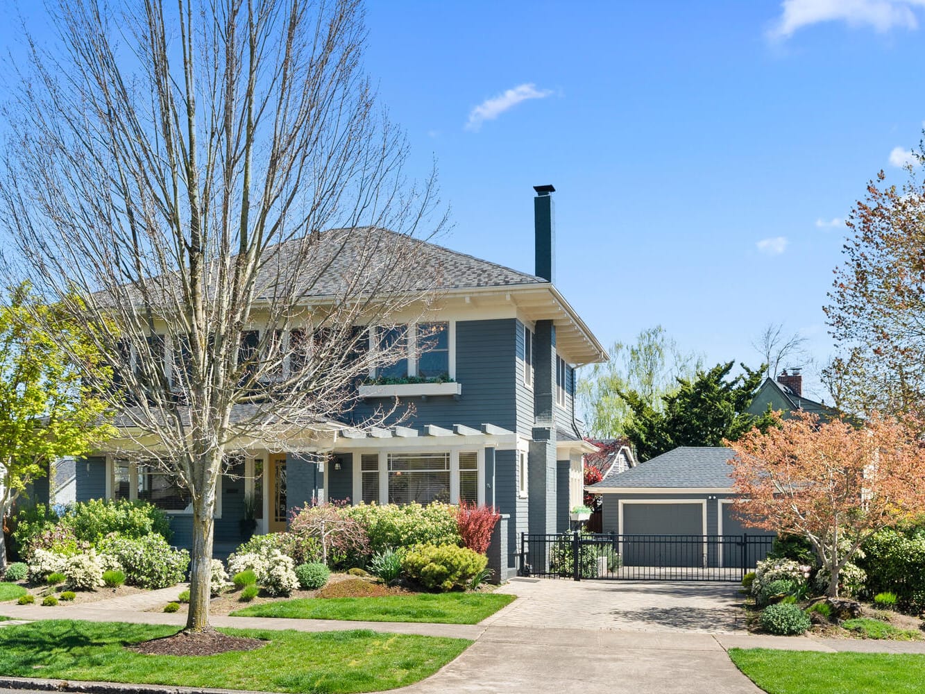 A two-story blue house with a pitched roof and chimney, surrounded by a manicured lawn and various trees and shrubs. A driveway leads to a detached garage. The sky is clear and blue.