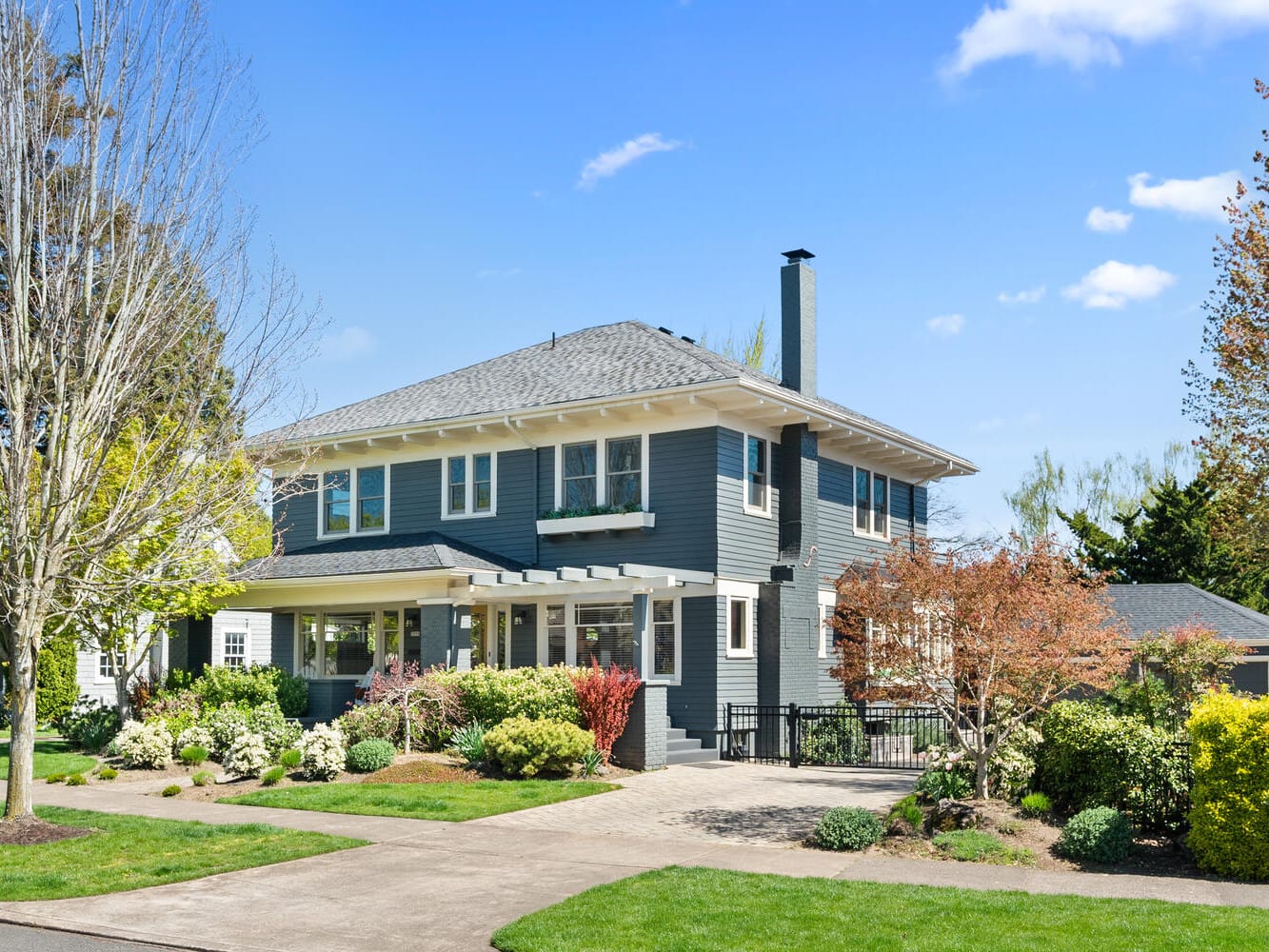 Large two-story gray house with white trim, surrounded by a landscaped garden, trees, and shrubs. A clear blue sky is in the background. The house features a chimney and a side garage.