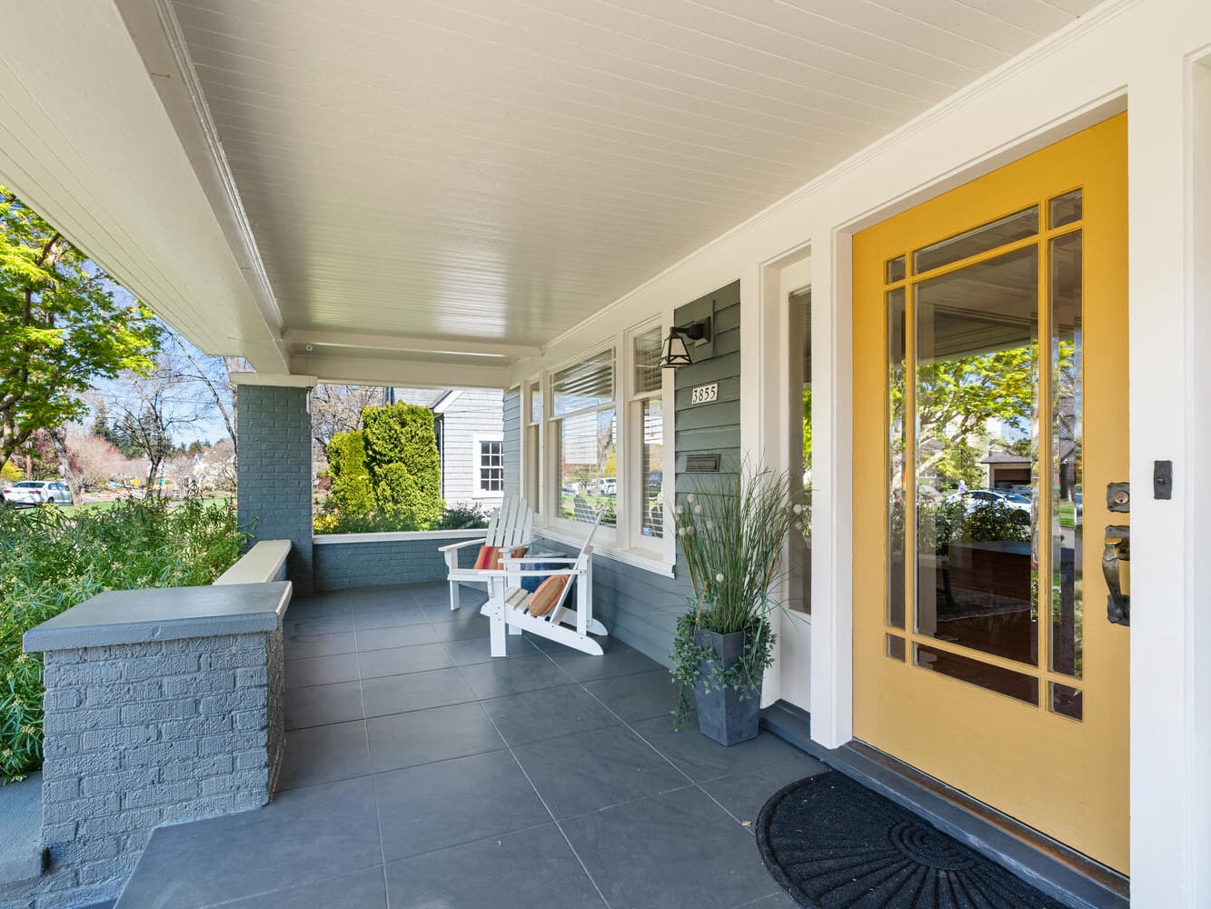 A charming porch with gray tiles and a bright yellow door. Two white chairs with colorful cushions are on the left, framed by green potted plants. The porch is shaded and overlooks a garden with trees, capturing a peaceful outdoor setting.