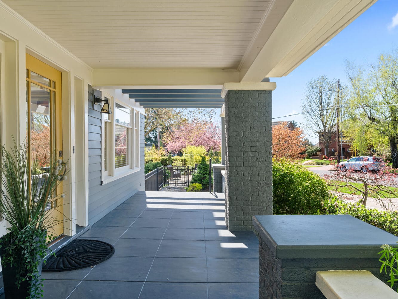 A sunny front porch with gray tiles, a wooden ceiling, and a yellow door. The porch is decorated with small plants, and theres a view of a garden with pink blossoms and a street lined with trees in the background.