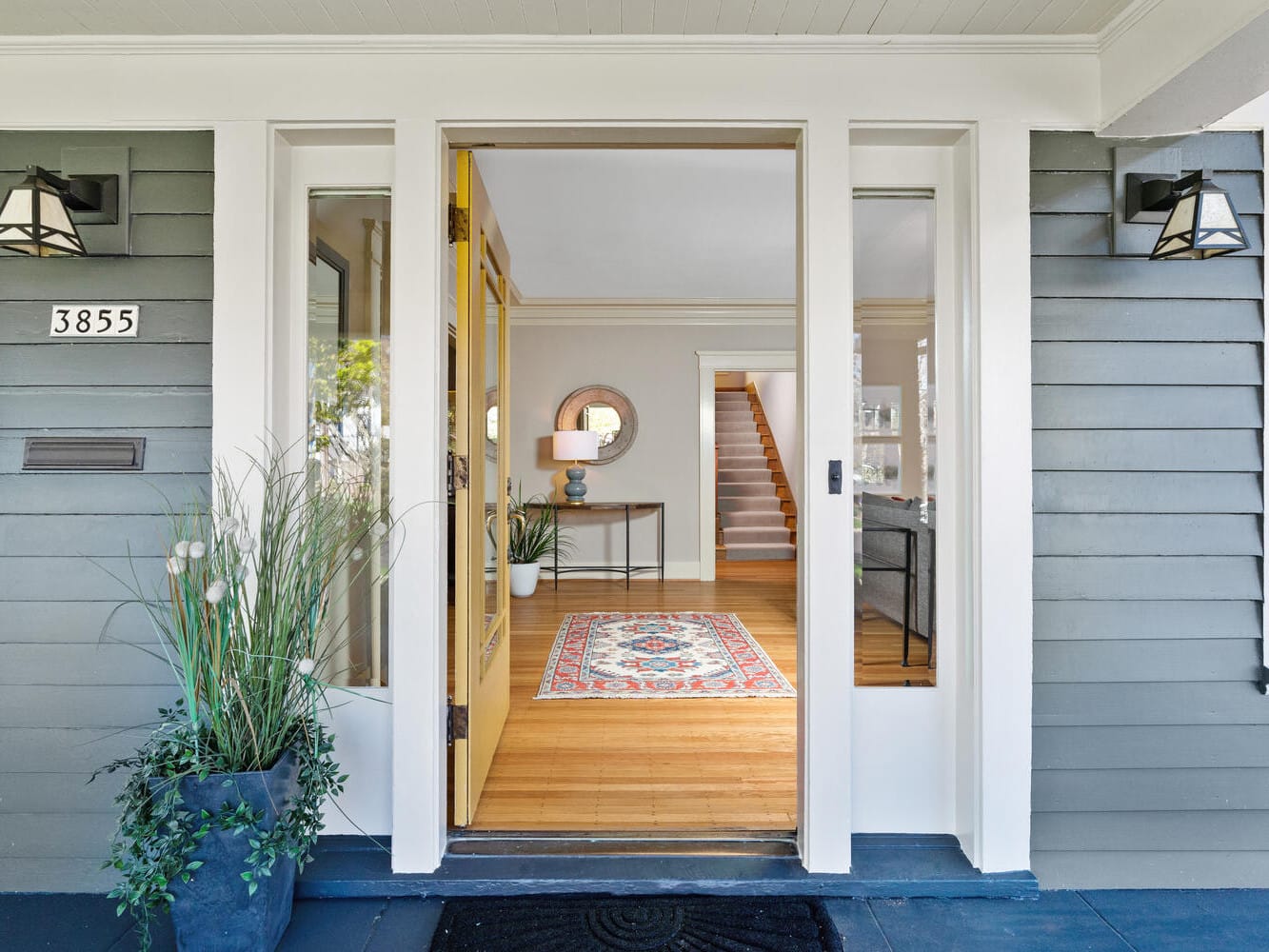 Front porch of a house with gray siding, featuring a welcoming open door. Inside, a hallway displays a colorful rug and round mirror. Potted plants flank the entryway, enhancing the homes inviting and modern ambiance.