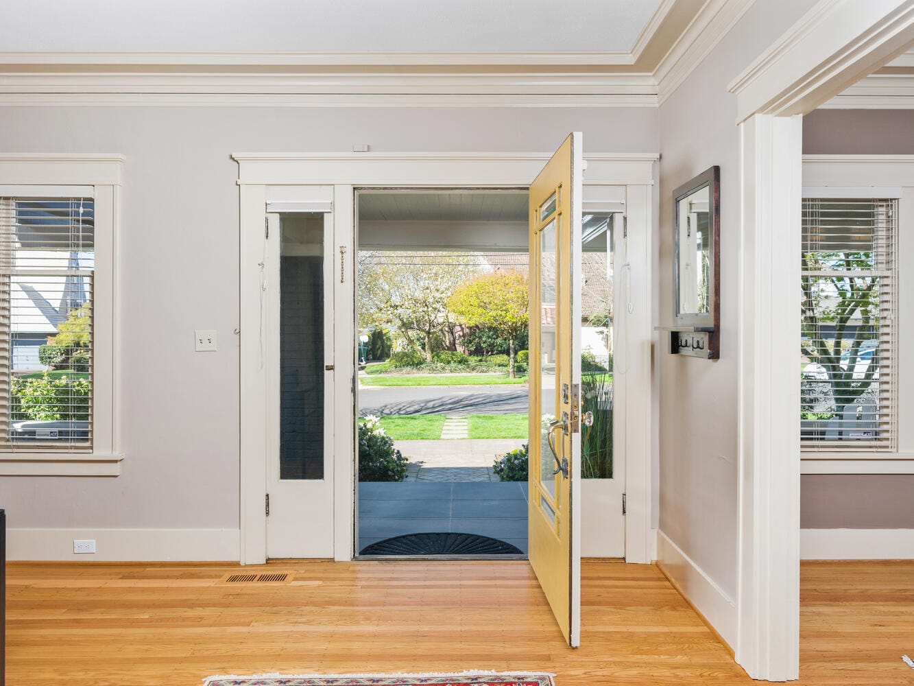 Open front door of a bright living room with hardwood floors, leading to a view of a sunny neighborhood. Large windows with blinds flank the door, and a small black doormat is on the porch outside.