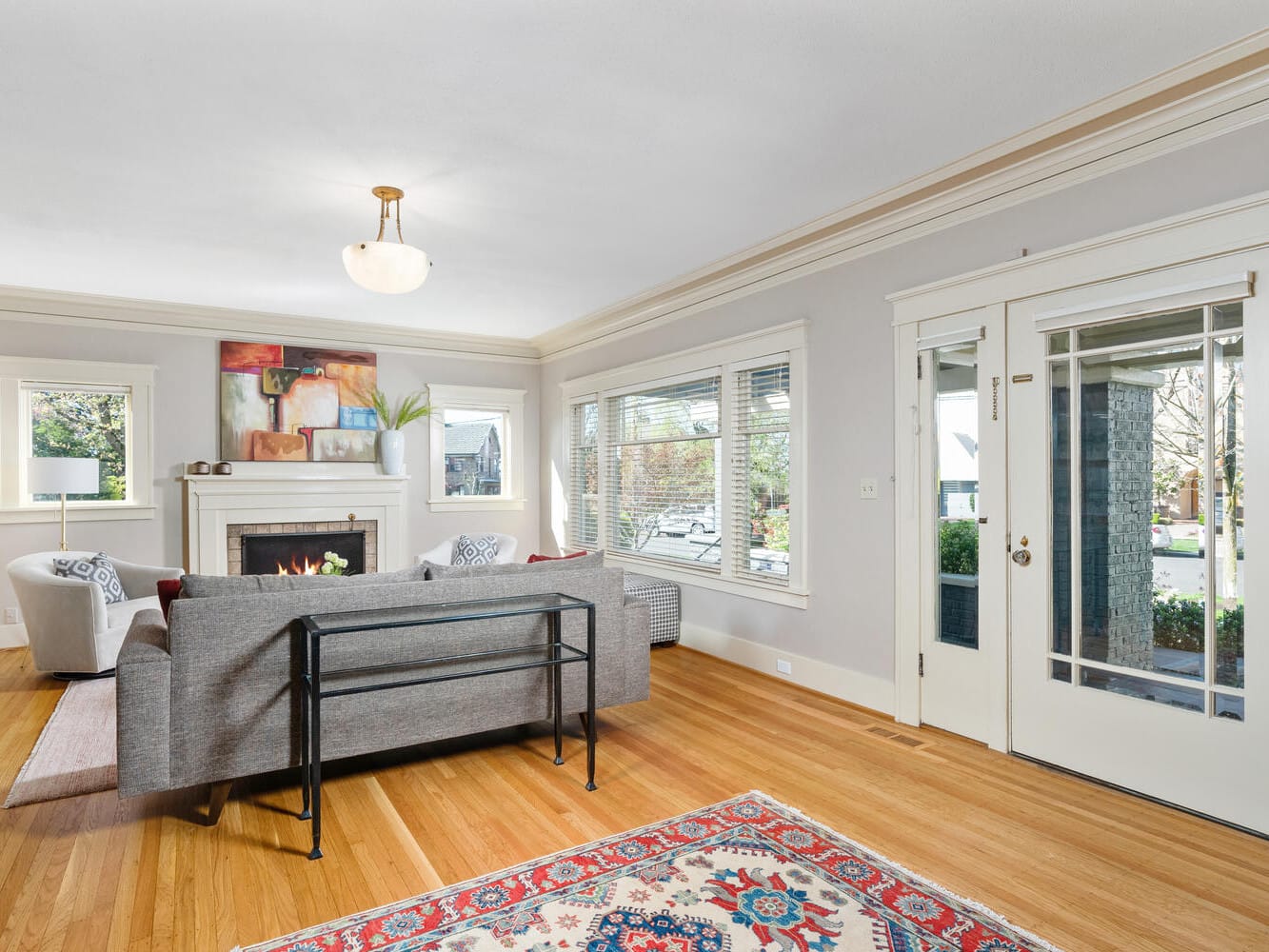 A bright living room with a gray sofa facing a fireplace, decorated with modern artwork. Large windows and a glass door let in natural light. A colorful rug covers the wooden floor, and two armchairs sit by the windows.