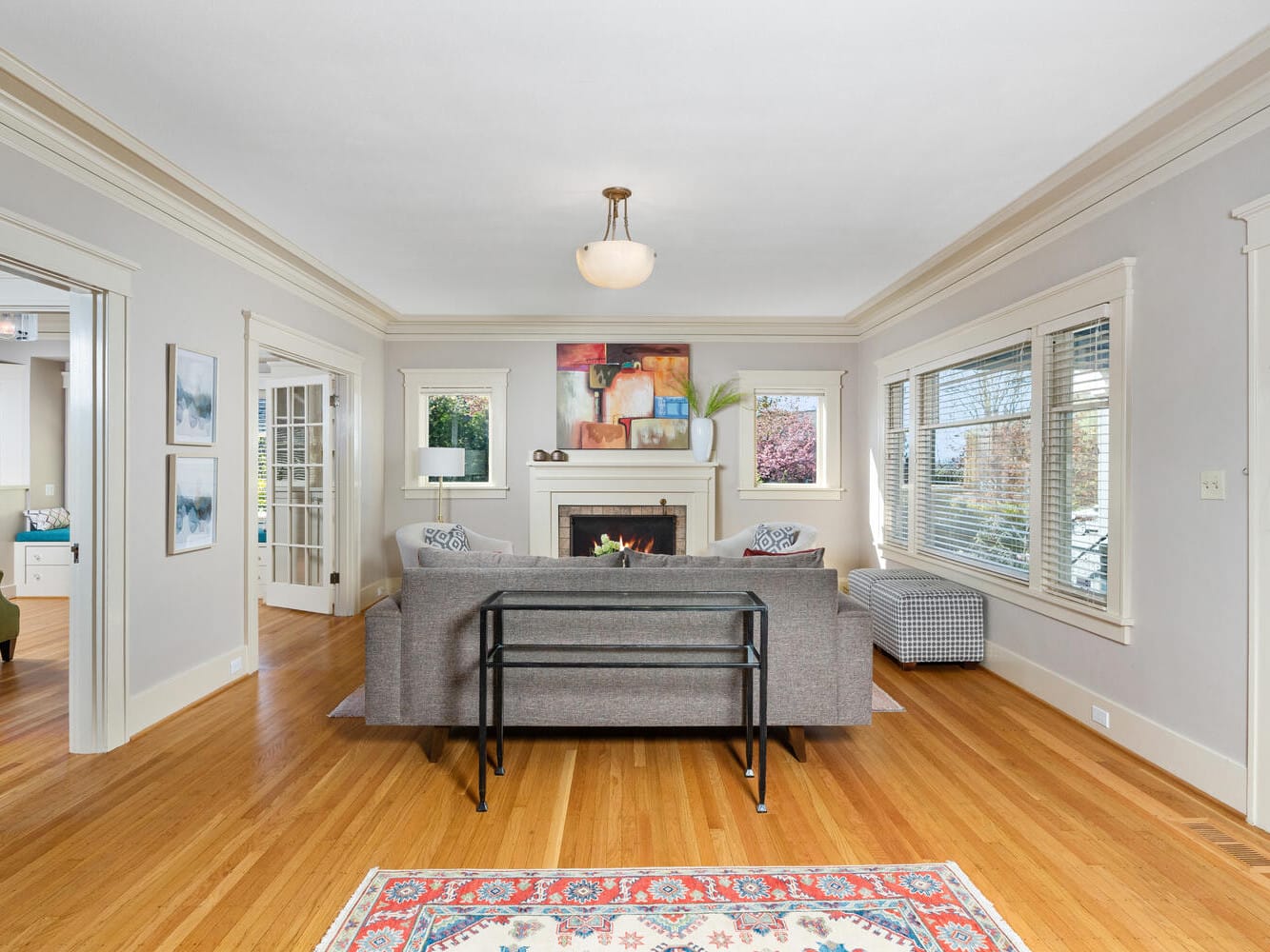 A cozy living room with hardwood floors, featuring a gray sofa facing a fireplace adorned with artwork. Large windows allow natural light to fill the space. White walls are decorated with framed pictures. An adjacent room with green chairs is partially visible.