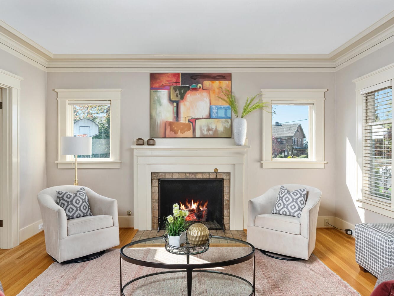 A cozy living room with a lit fireplace, flanked by two beige armchairs with patterned cushions. A round coffee table in the center, under a colorful abstract painting. Two windows with blinds allow natural light in, highlighting the warm wood floor.