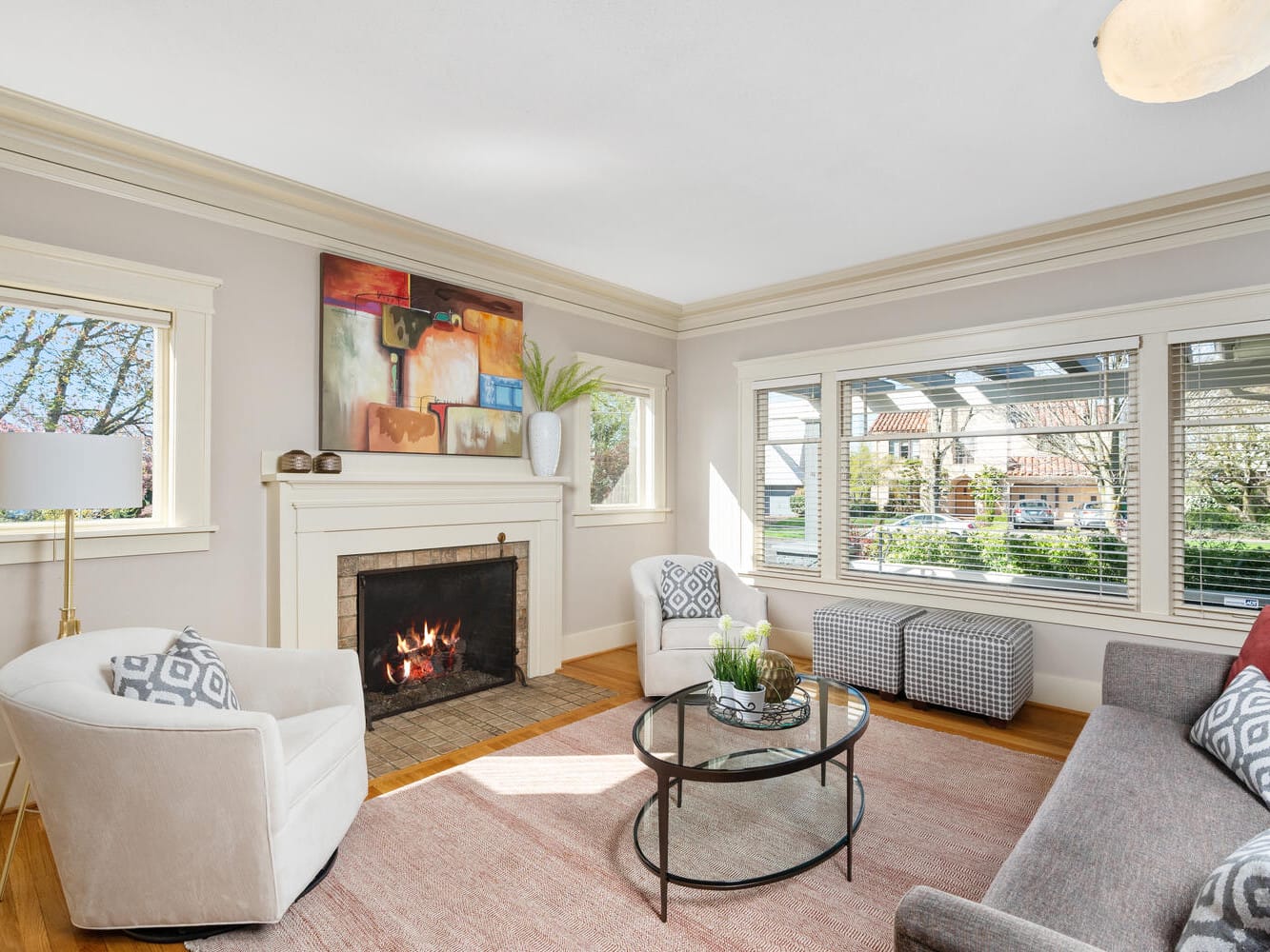 A bright living room with a fireplace, modern art above it, and a cozy fire. The neutral-toned room features a gray sofa, a white armchair, a glass coffee table, and large windows that let in natural light. A red pillow and decorative plants add color.