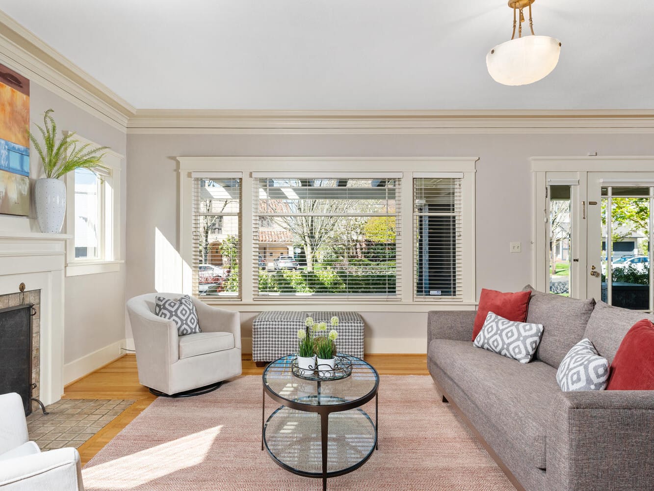 A bright living room with a large window, a gray sofa with red and patterned cushions, a beige armchair, and a round glass coffee table. A fireplace on the left features modern art and a decorative vase. Light wooden floors add warmth.