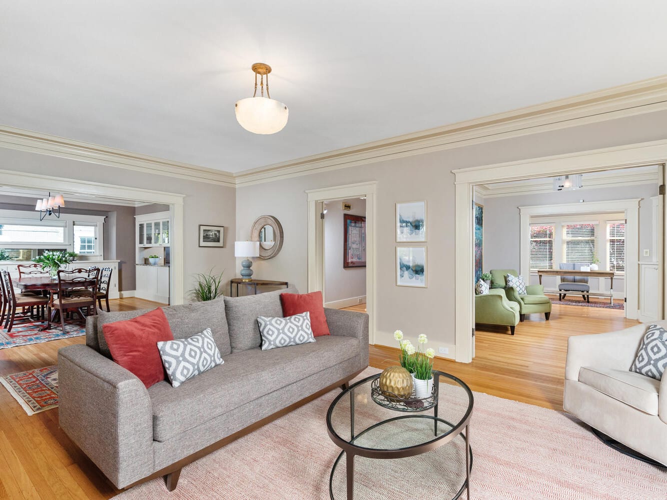 A spacious living room featuring a gray sofa with patterned pillows, a glass coffee table, and a cozy armchair. The view extends to a dining area with a wooden table and chairs. Wood flooring and soft lighting create a warm atmosphere.