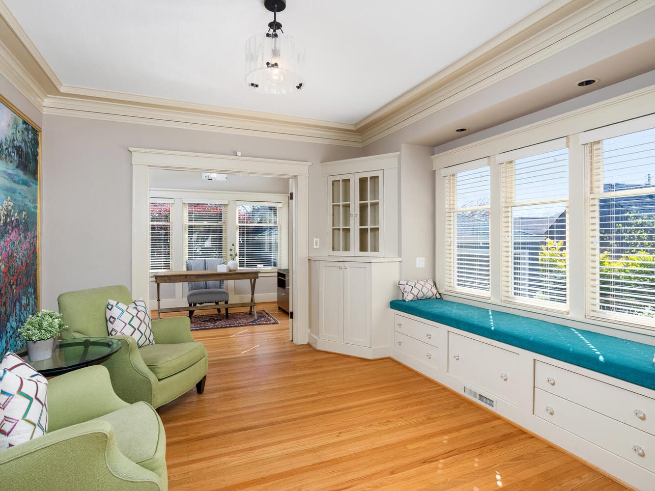 A cozy, sunlit room featuring two green armchairs, a round glass table, and a large bay window with turquoise cushions and storage drawers. A colorful painting adorns the wall, and an adjoining office area is visible through an open door.