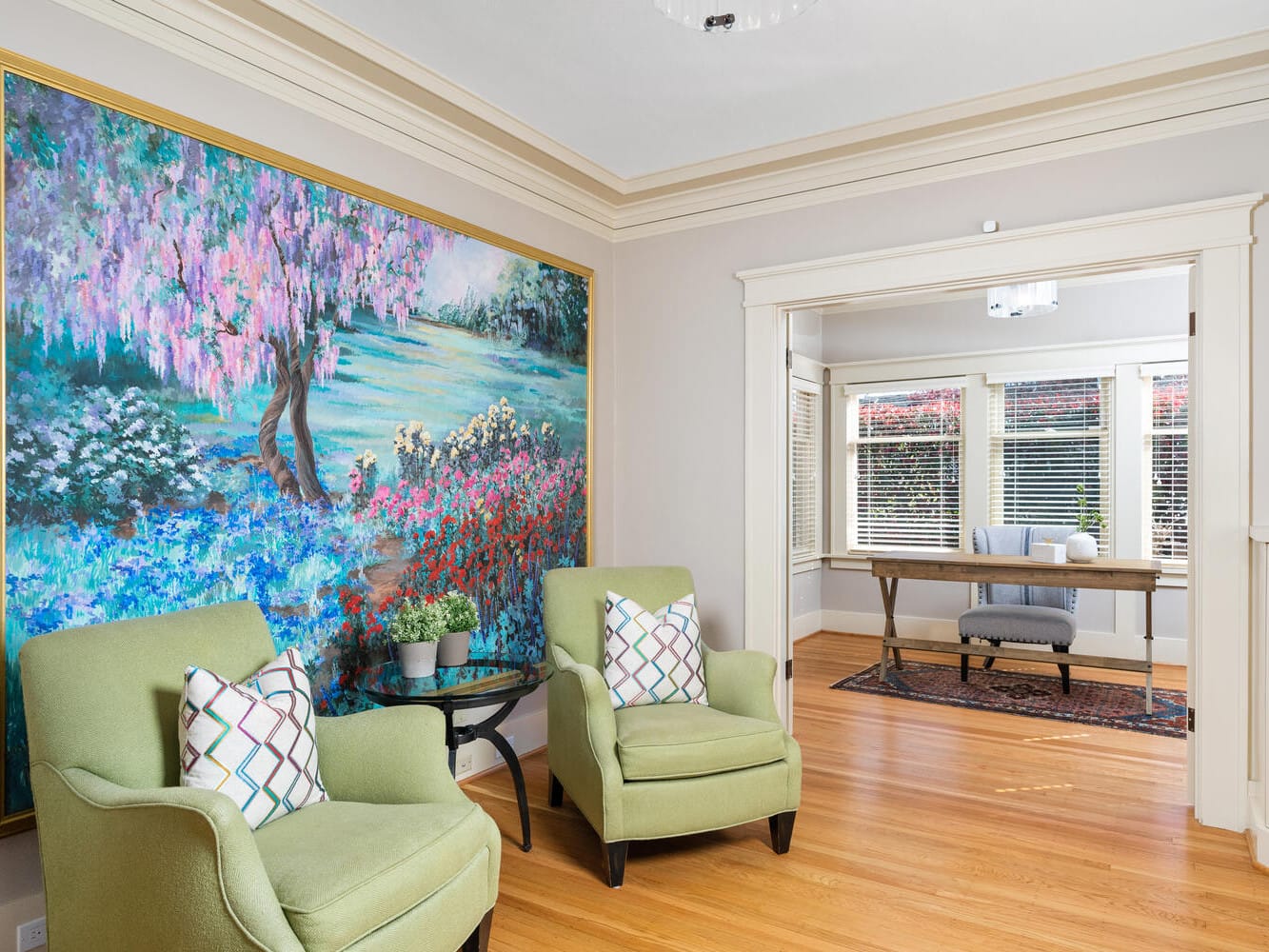 A bright living room with two green armchairs and colorful throw pillows. A large, vibrant painting of a floral landscape hangs on the wall. A wooden floor extends to a dining area with a table and bench, surrounded by windows with blinds.