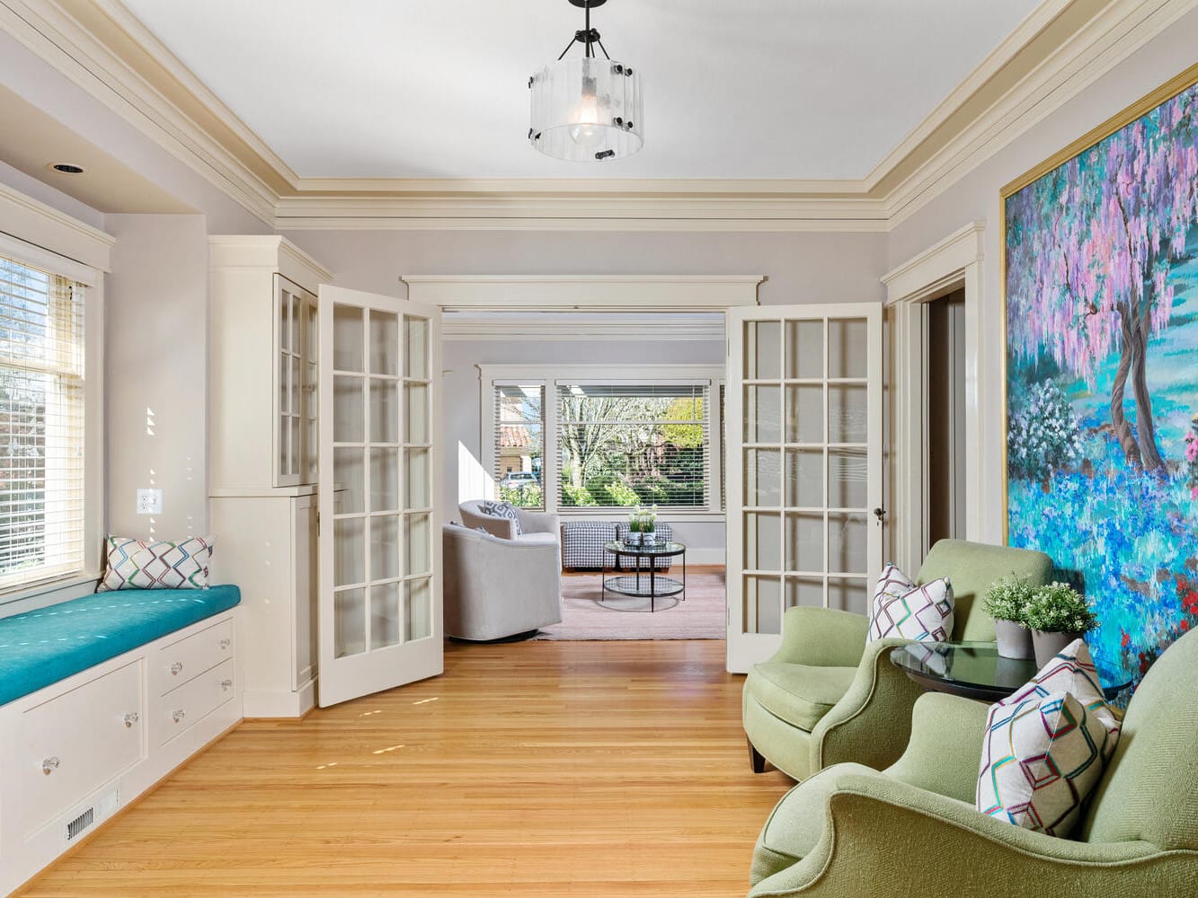 A bright, spacious living room with light wooden floors, green armchairs, a colorful painting on the wall, and glass-paneled doors opening to another room. A window seat with blue cushions is on the left, overlooking a sunlit garden.