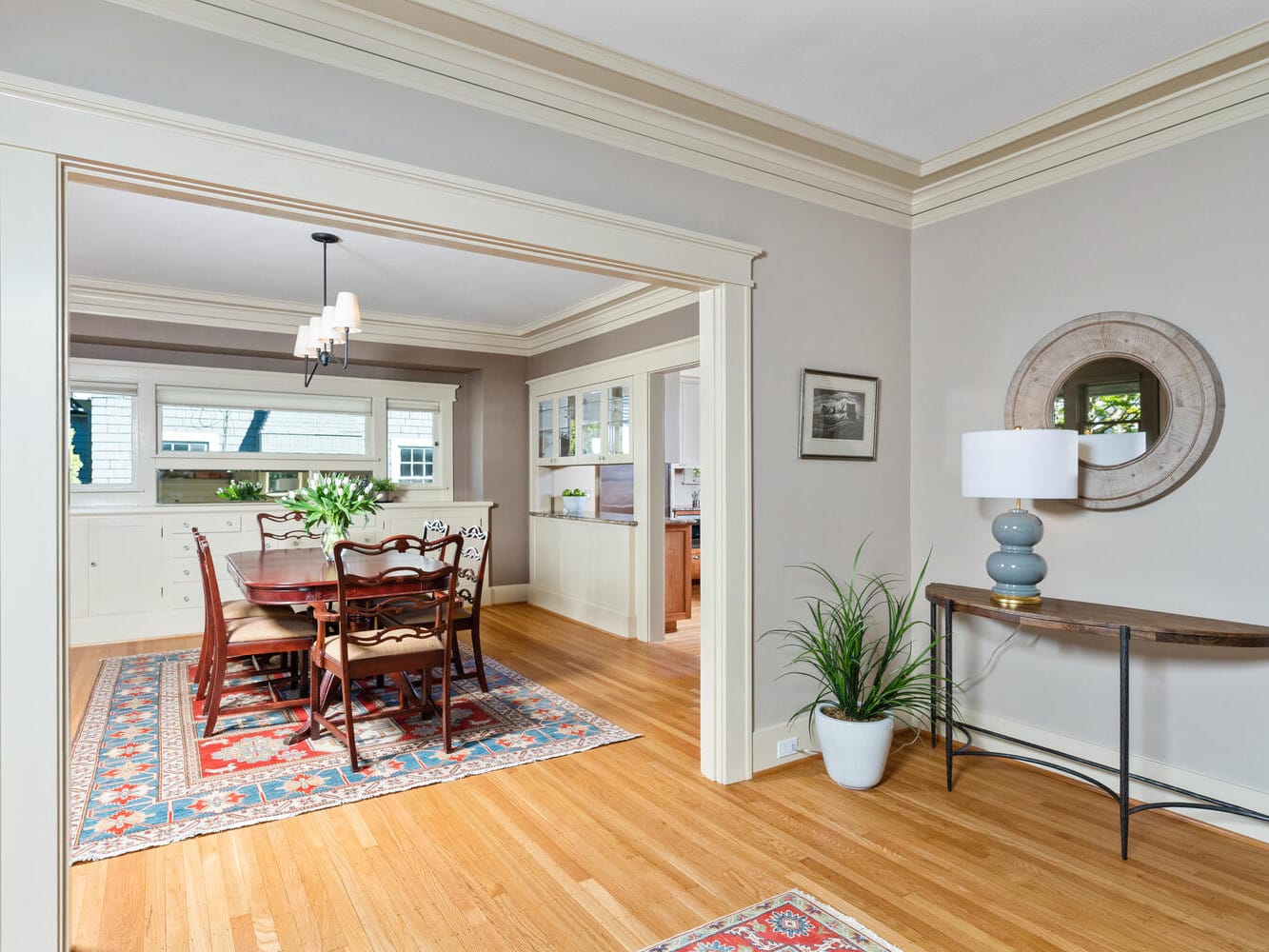 A dining room with a wooden table and chairs, a patterned rug, and a sideboard beneath large windows. A round mirror and lamp sit on a console table nearby, alongside a potted plant on the wooden floor. The walls are painted light gray.