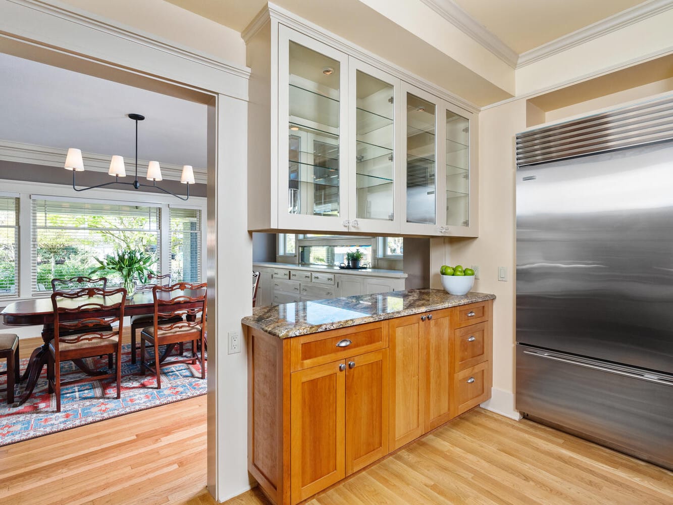 A modern kitchen featuring wooden cabinets, a granite countertop, a large stainless steel refrigerator, and glass-fronted upper cabinets. In the background, theres a dining room with a wooden table and chairs, under a chandelier.