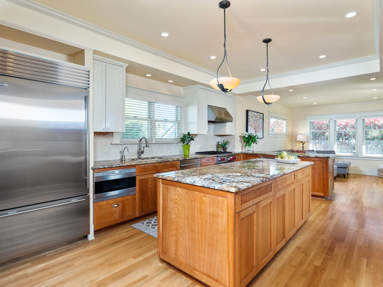 A spacious kitchen with wooden flooring and a large island. It features stainless steel appliances, including a refrigerator and oven, granite countertops, pendant lights, and bright windows. Green plants and fruit bowls add a touch of color.