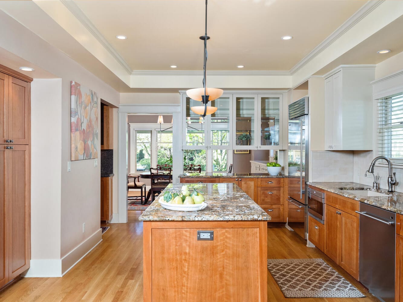 A spacious kitchen with wooden cabinets, a large island with a granite countertop, and a double sink. Pendant lights hang above the island, and a fruit bowl is on top. The room has hardwood flooring and large windows letting in natural light.