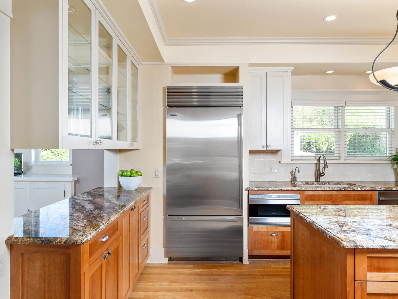 A modern kitchen with wooden cabinets and granite countertops. A stainless steel refrigerator stands between two sets of cabinets. A sink with a faucet is near a window, and a bowl of green apples is on the counter. A hanging light fixture is above.