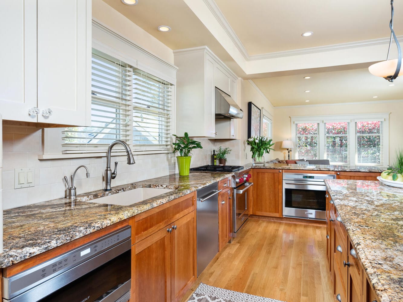 A spacious kitchen with granite countertops, wooden cabinets, and stainless steel appliances. It features a central island, a sink with a window view, and potted plants for decoration. The space is well-lit with overhead lighting.