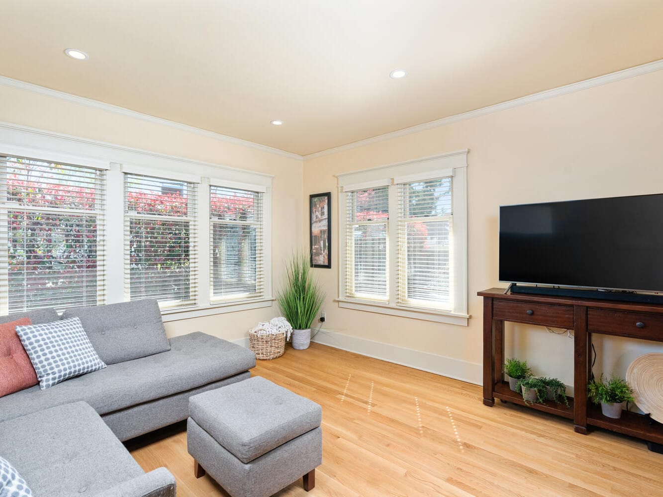 A cozy living room with a gray sectional sofa and an ottoman. A flat-screen TV sits on a wooden stand. Large windows with blinds let in natural light. The room is decorated with plants and a basket, and has light-colored walls and a wooden floor.