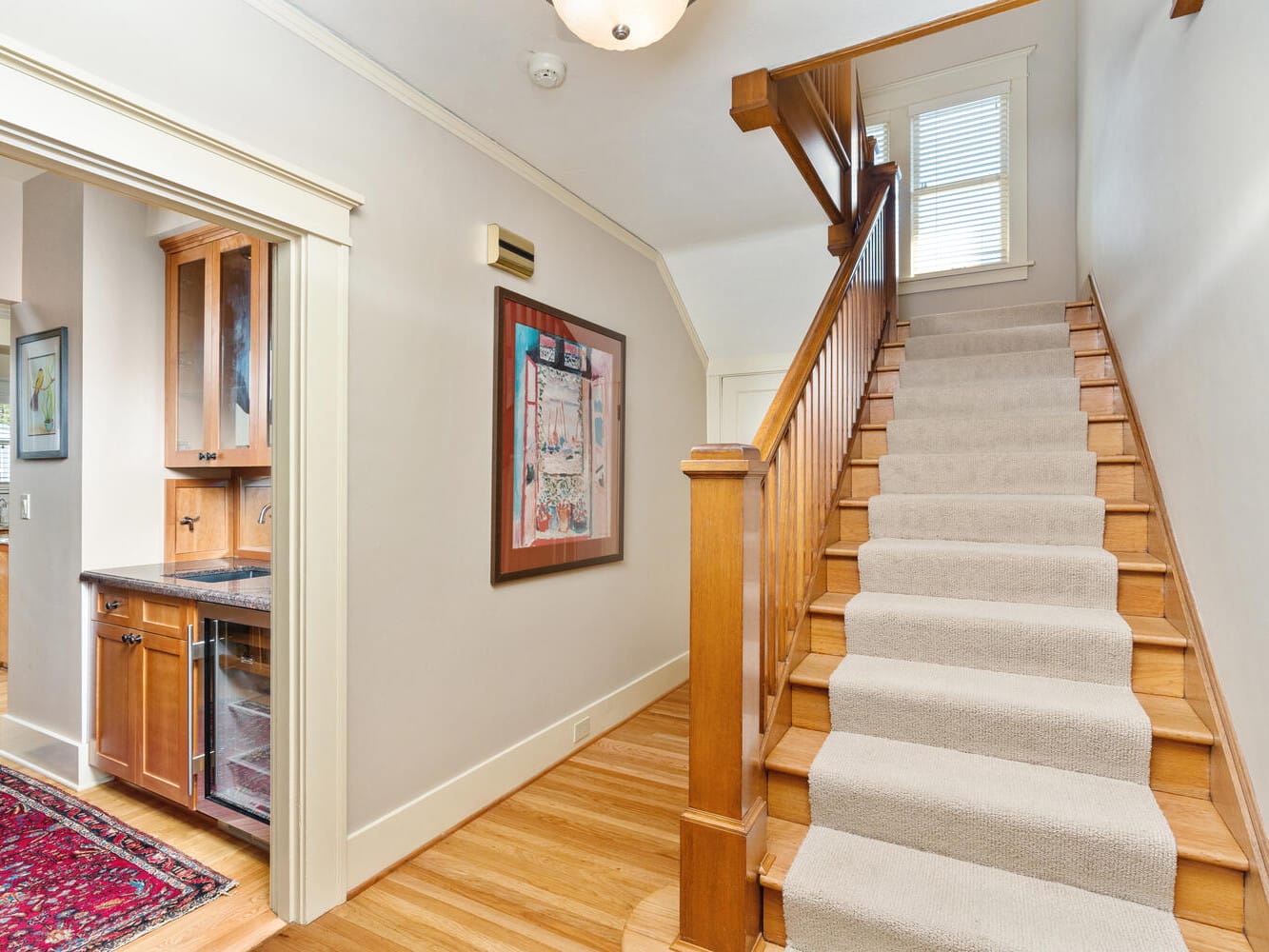 A hallway features a staircase with a wooden banister and carpeted steps leading upwards. On the left, theres an open doorway to a kitchen with wooden cabinets and a small red rug. A colorful framed artwork hangs on the wall.