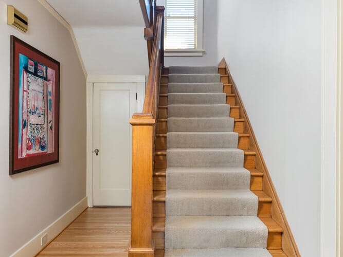 A hallway with wooden floors features a staircase with beige carpet and wooden bannister leading upwards. A closed white door sits beneath the stairs, and a colorful painting hangs on the left wall. A ceiling light fixture is above.