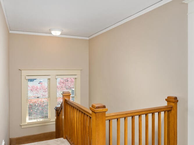 Wooden staircase with beige carpet, leading to a window with closed blinds. Walls are painted light beige, and a ceiling light illuminates the space.