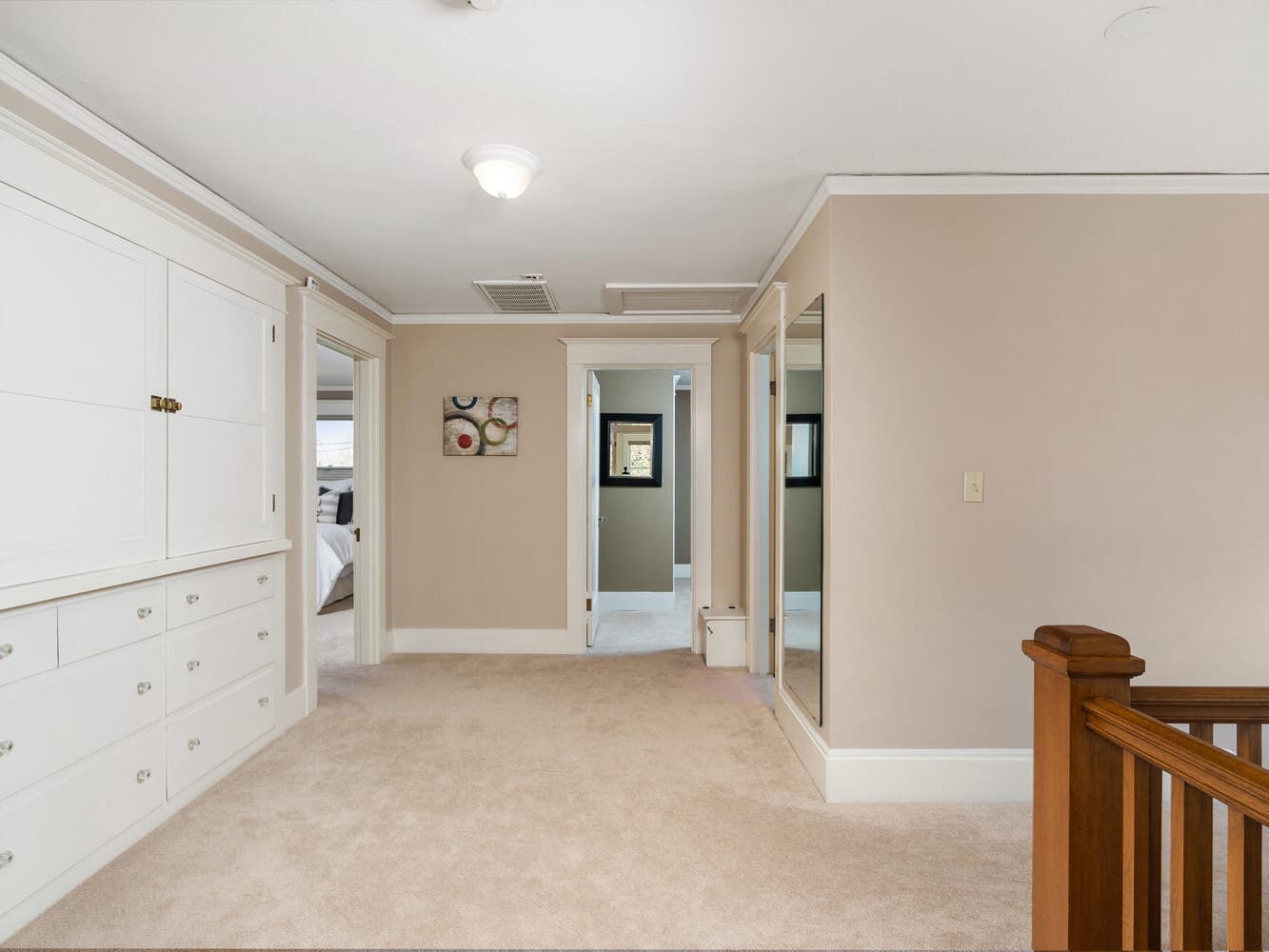 A well-lit hallway with beige walls, carpeted floor, and built-in white cabinets. A wooden stair railing is on the right. Doors lead to bedrooms, and a framed picture hangs on the wall. A ceiling light illuminates the space.