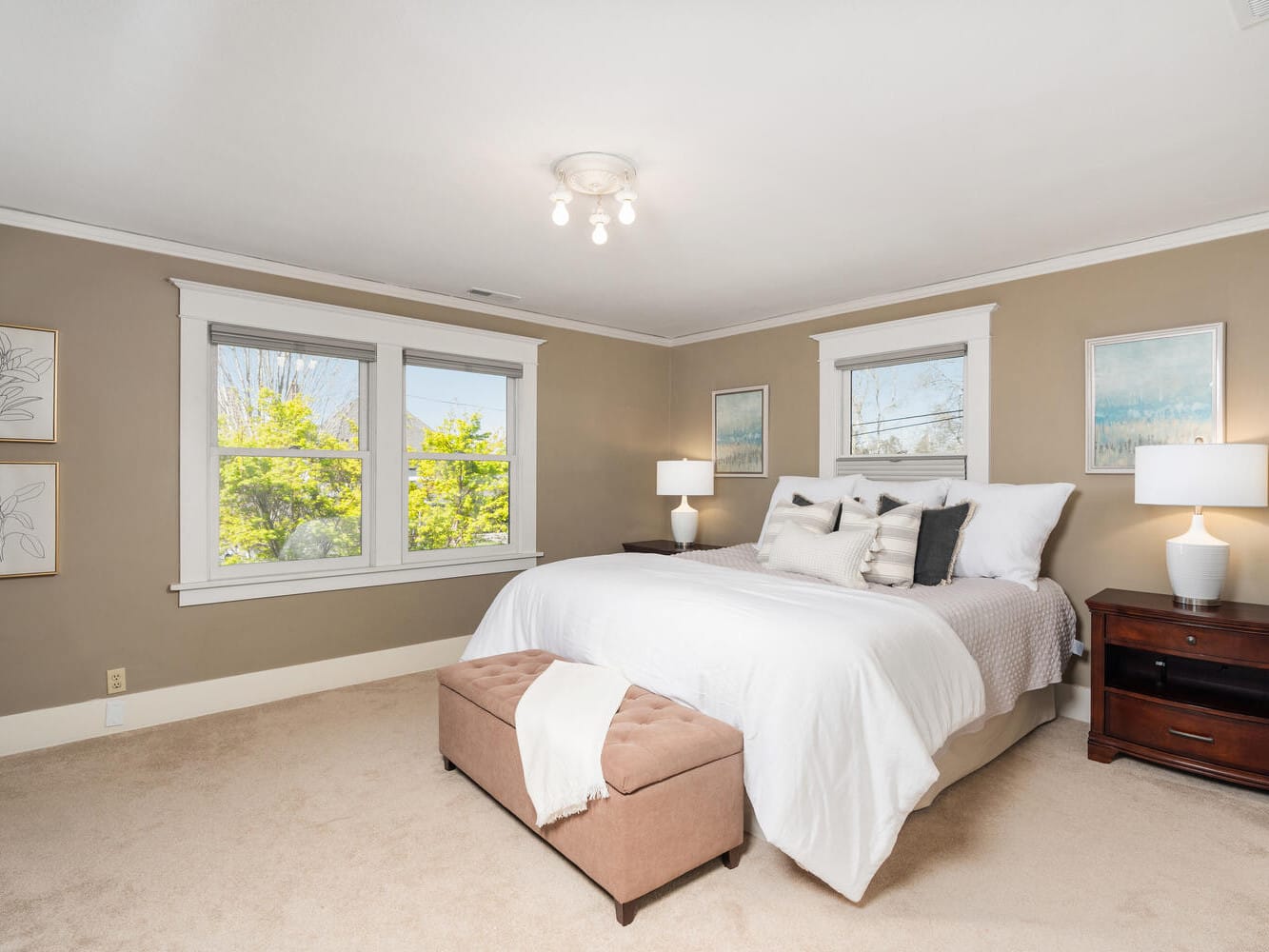 A cozy bedroom with beige walls features a large bed with white bedding and multiple pillows. There are two wooden nightstands with lamps on either side of the bed. A soft brown ottoman sits at the foot of the bed. Two windows let in natural light.