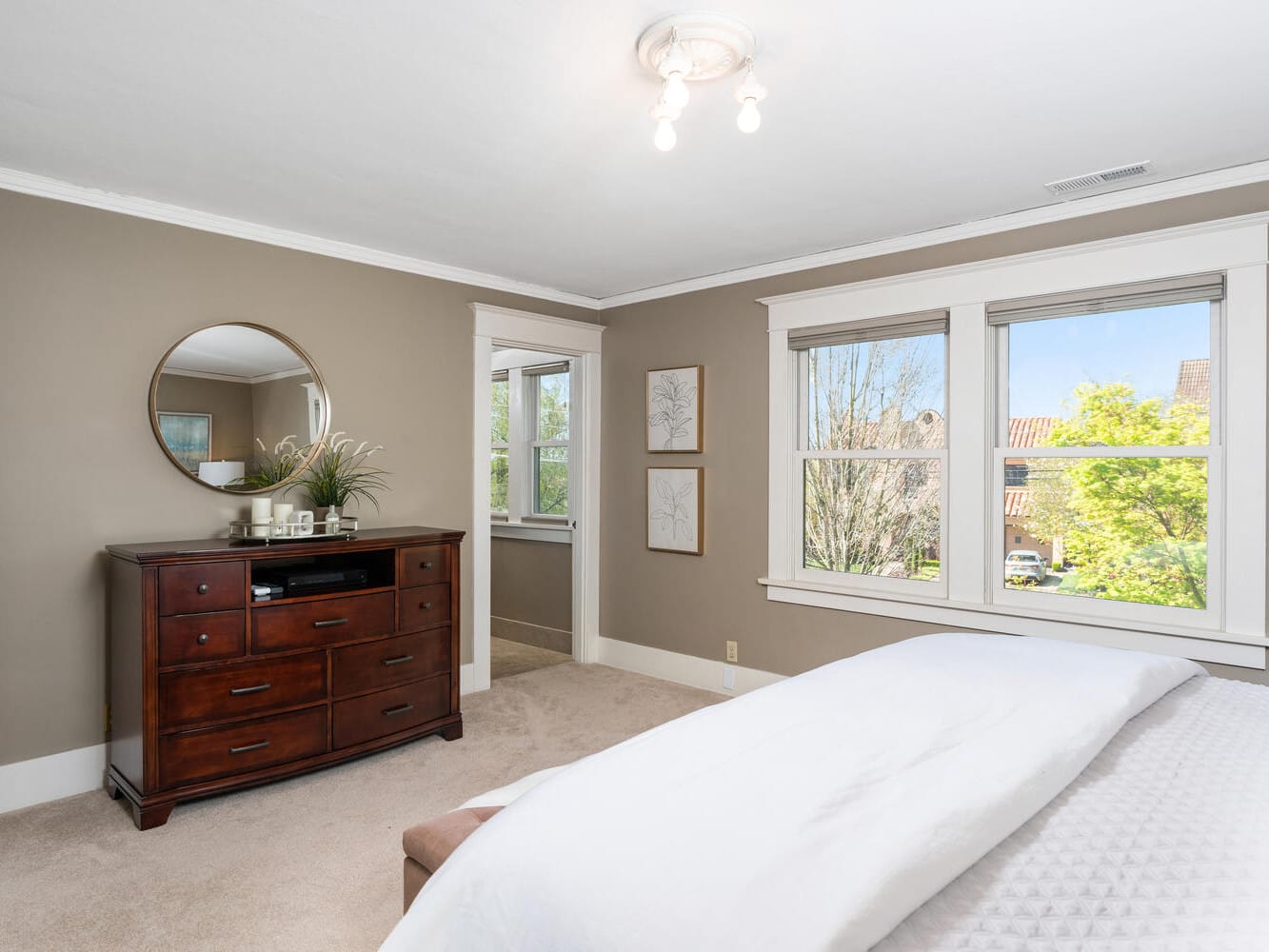 A cozy bedroom featuring a wooden dresser with a round mirror above it, artwork on the walls, a bed with white bedding, and large windows allowing natural light. The room has beige walls and carpeted floors.