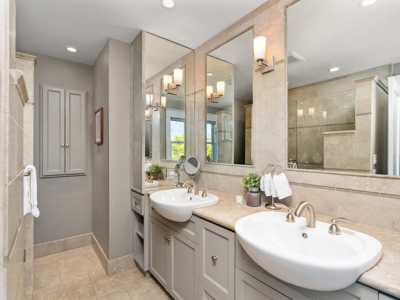 A modern bathroom with two white sinks on a beige countertop, large mirrors, wall-mounted lights, and light gray cabinets. A towel hangs near a walk-in shower, and two small potted plants add greenery.