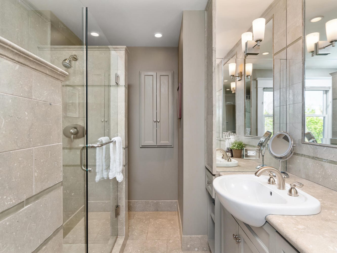 A modern bathroom featuring a glass-enclosed shower, a sink with a large mirror, and elegant lighting. The walls and floor are tiled in neutral stone tones, creating a sleek and inviting atmosphere.