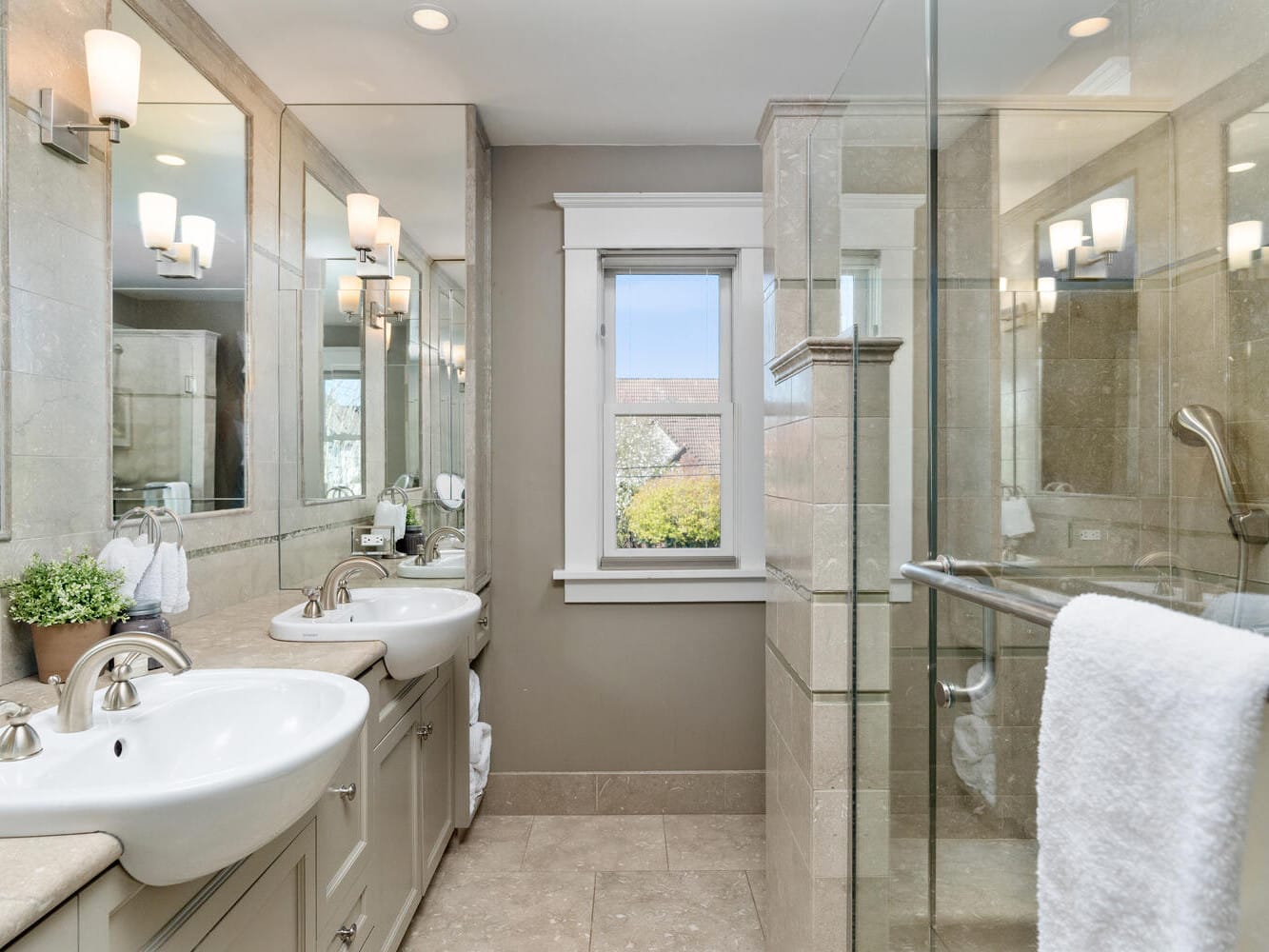 A modern bathroom with two sinks, a large mirror, and wall-mounted lights. There is a glass-enclosed shower on the right, a small window in the center, and a potted plant on the counter.