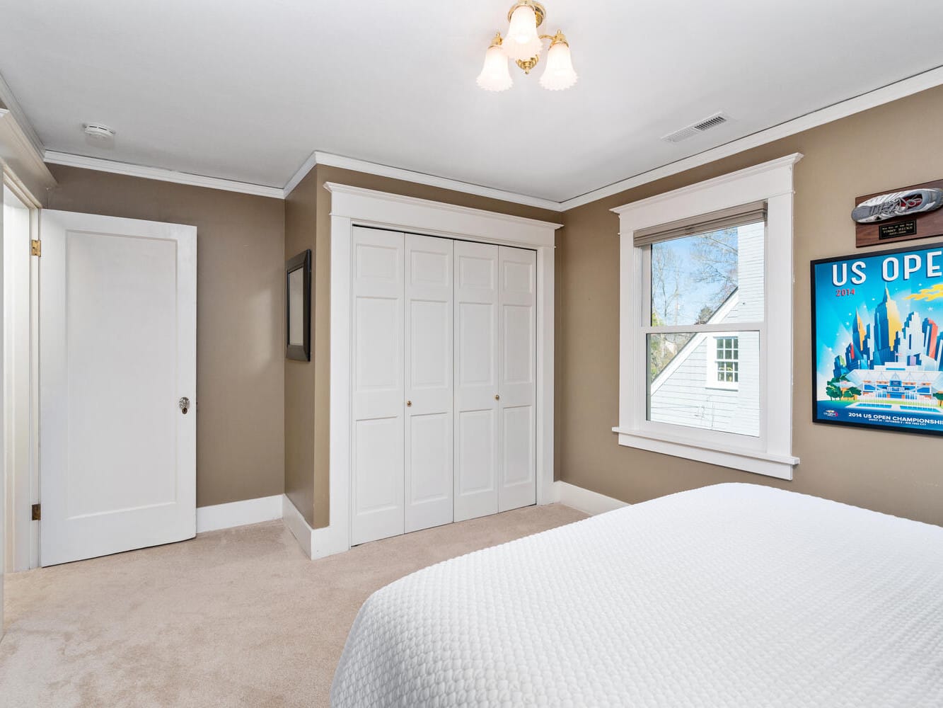 A bedroom with beige walls and carpet, featuring a white bedspread. Theres a window with a view of a roof, a closed white closet, and a poster on the wall. A ceiling light fixture is centered above.