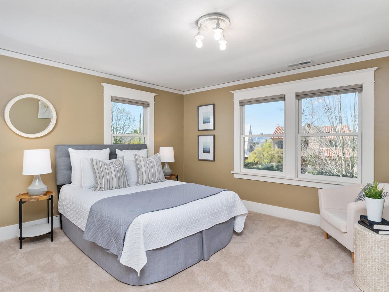 A cozy bedroom features a neatly made double bed with gray and white bedding, flanked by two wooden nightstands with lamps. A round mirror and framed artwork hang on beige walls. Large windows allow natural light in, and there’s an armchair with a plant.