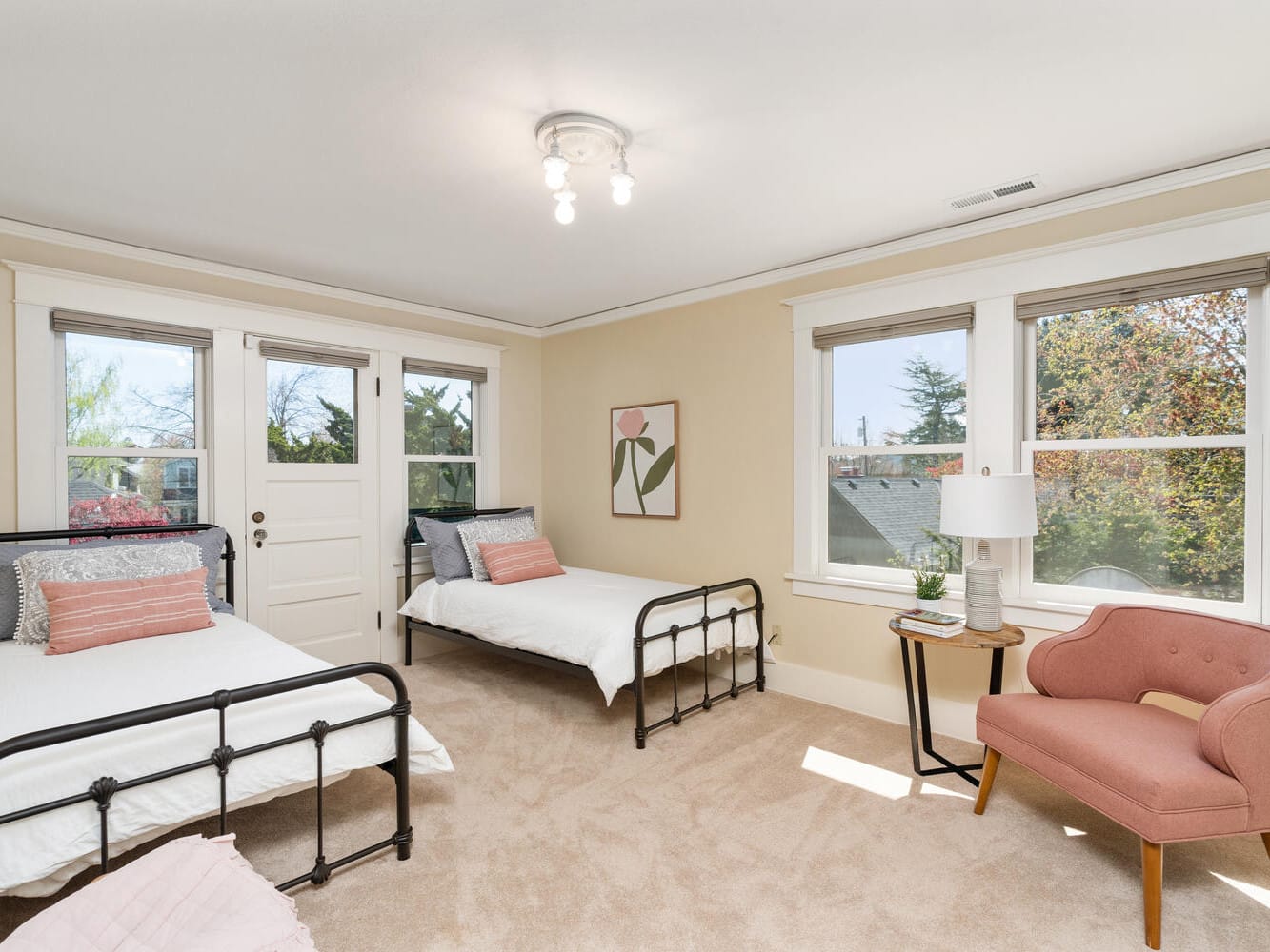 A bright bedroom with two metal-frame twin beds, a pink accent chair, and a small table with a lamp. Large windows let in natural light, and a painting of a tulip hangs on the wall. The walls are cream-colored, and the floor is carpeted.